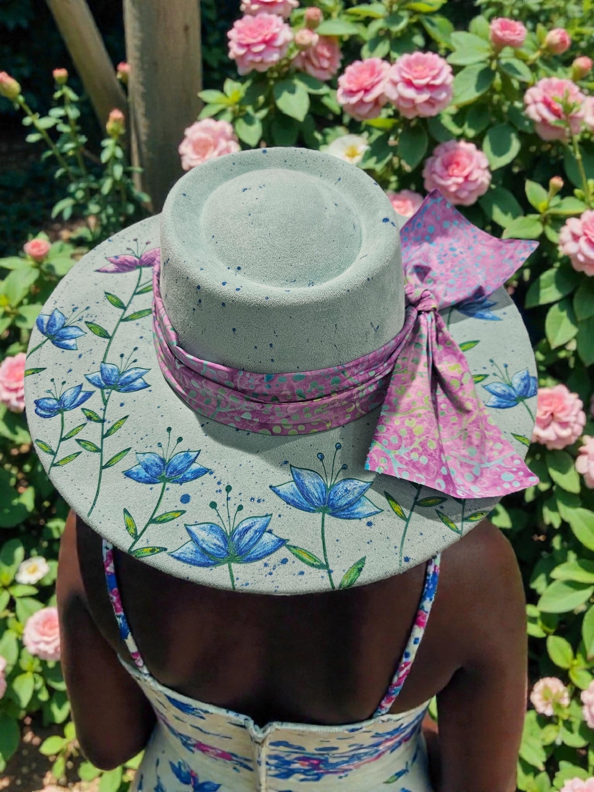 Person wearing hand-painted sparkle and laser-engraved vegan suede rancher hat with batik band, hat features a floral pattern design. In a garden setting; back view showing custom artwork