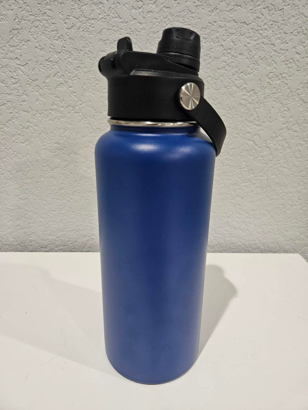 backside of a 32oz Blue hydro water bottle with black lid on a white surface against a gray wall