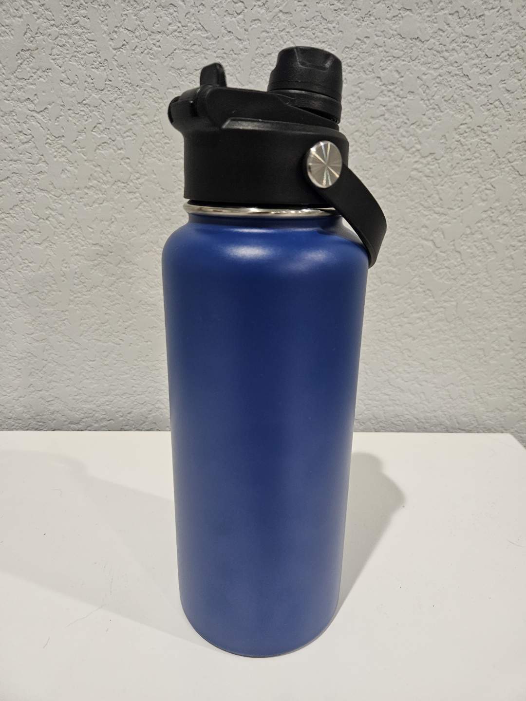 backside of a 32oz Blue hydro water bottle with black lid on a white surface against a gray wall
